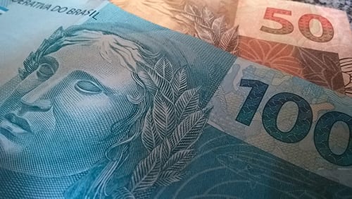 A imagem mostra uma nota de cem reais em cima de uma nota de cinquenta reais.