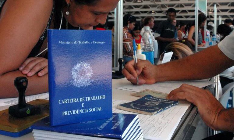 Imagem de Carteira de Trabalho em pé enquanto pessoa assina contrato. Caged: Brasil tem saldo de 328 mil empregos em fevereiro