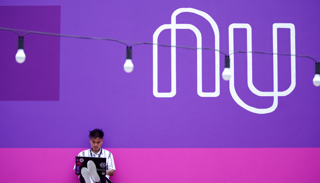 Imagem mostra um jovem trabalhando com um painel do Nubank de fundo.