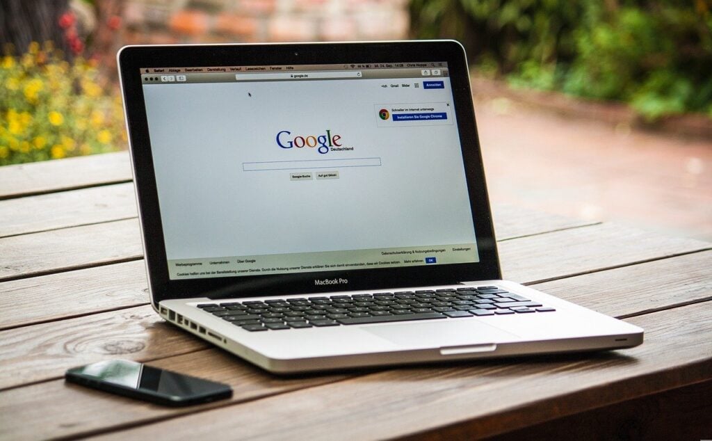 Laptop aberto sobre uma mesa tendo um aparelho celular ao lado, e aberto na página do Google
