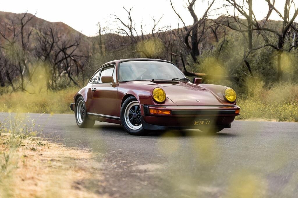 Modelo Posrche 911, na cor vinho, em uma estrada perto de uma floresta