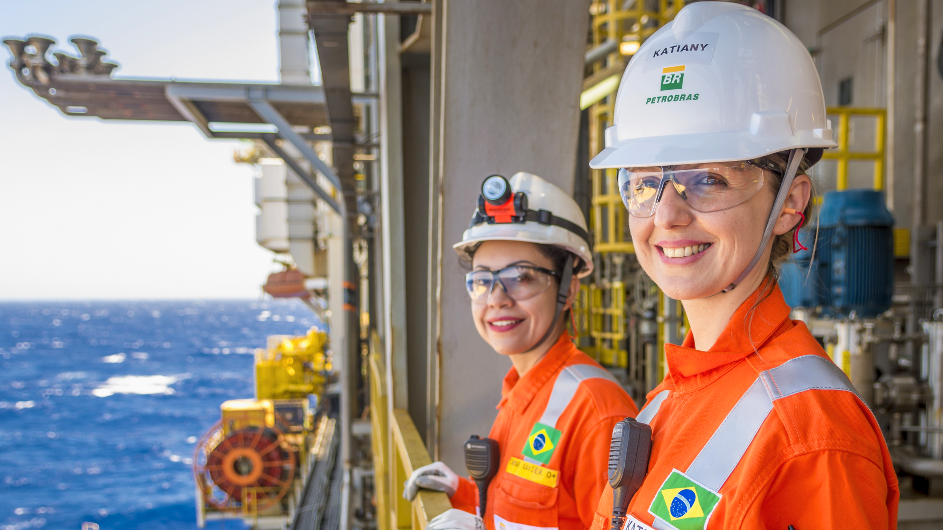 Duas profissionais em um ambiente industrial em uma plataforma de petróleo no mar. Elas vestem uniformes de segurança laranja com faixas reflexivas, capacetes de proteção com o logotipo da Petrobras e óculos de proteção.