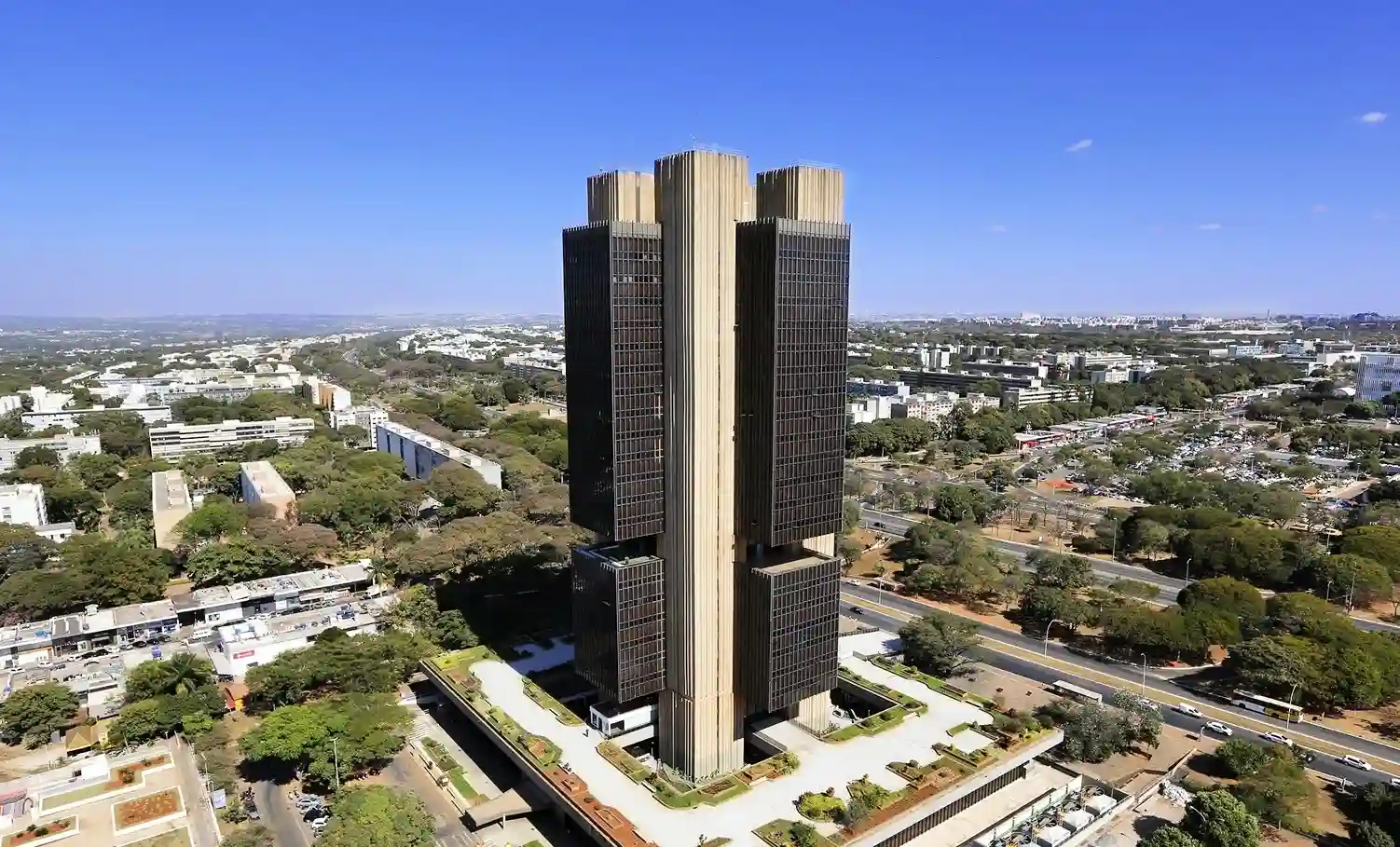 Vista aérea do edifício-sede do Banco Central do Brasil em Brasília, rodeado por áreas arborizadas e vias urbanas.