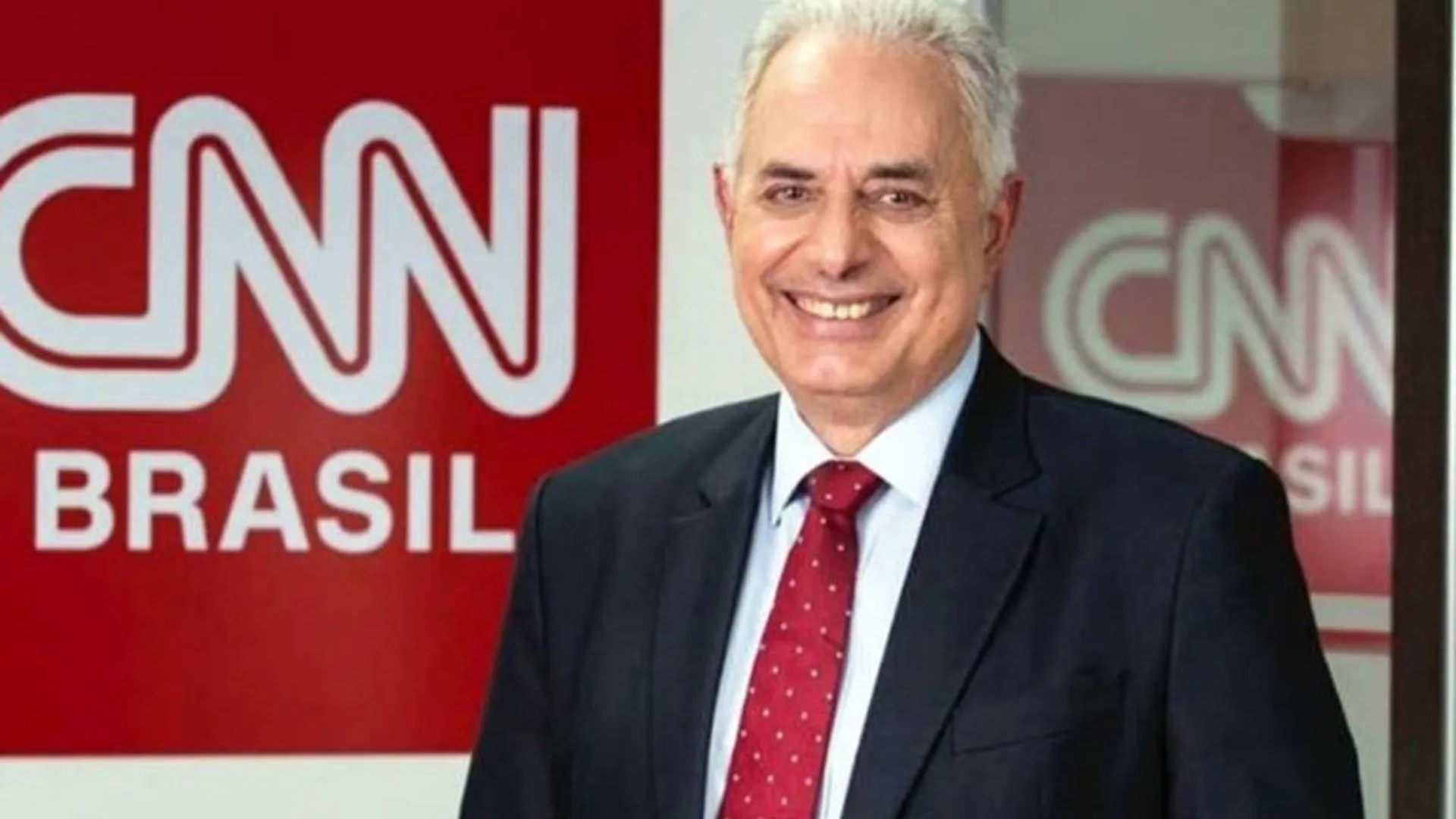 William Waack em frente ao logo da CNN Brasil, sorrindo e usando terno escuro com gravata vermelha.