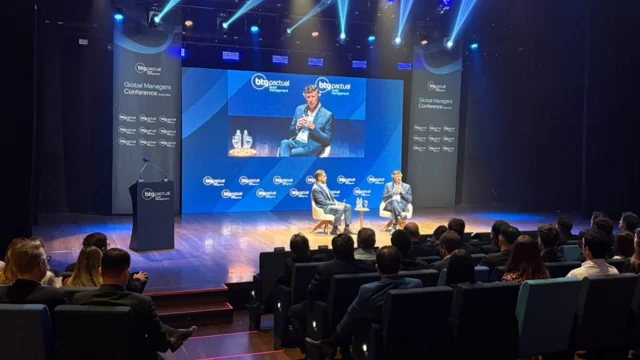 André Esteves e Mansueto Almeida sentados em poltronas brancas em um palco com painéis azuis da BTG Pactual Asset Management, participando de um debate sobre investimentos.