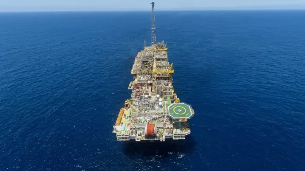Vista aérea de topo de uma grande plataforma de petróleo do tipo FPSO operando em alto-mar no oceano de águas azuis profundas. Em destaque na parte inferior da estrutura, observa-se o heliponto octogonal verde e amarelo com a letra "H" pintada, acompanhado de módulos industriais, tubulações de processamento e a torre de flare no topo da embarcação.