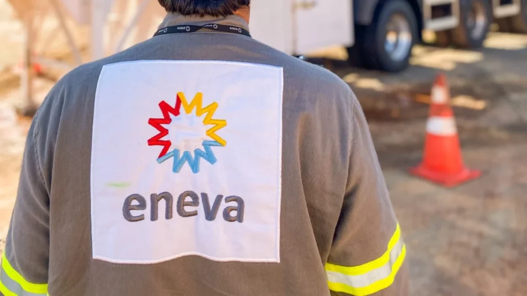 Fotografia em plano médio focado nas costas de um colaborador da Eneva vestindo jaqueta cinza de uniforme operacional com faixas refletivas amarelas e verdes nas mangas. No centro do casaco, destaca-se um patch quadrado de tecido branco com o logotipo colorido da Eneva em formato de estrela/sol nas cores vermelha, amarela e azul, com a marca "eneva" gravada em cinza-escuro. Ao fundo, com profundidade de campo desfocada, avistam-se as rodas de um veículo pesado e um cone de tráfego laranja e branco em área industrial.