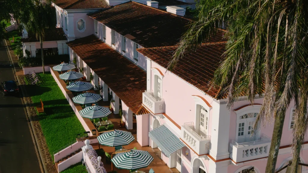 Vista aérea da fachada rosa do Hotel das Cataratas, com arquitetura colonial portuguesa, telhados de cerâmica, varandas com guarda-sóis listrados e jardins cercados pela Mata Atlântica